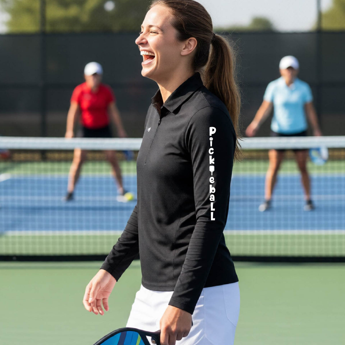 A woman wearing a black quarter-zip long-sleeve t-shirt with a white graphic text 'PICKLEBALL' on the sleeve with a pickleball paddle replacing the L, standing on a pickleball court.