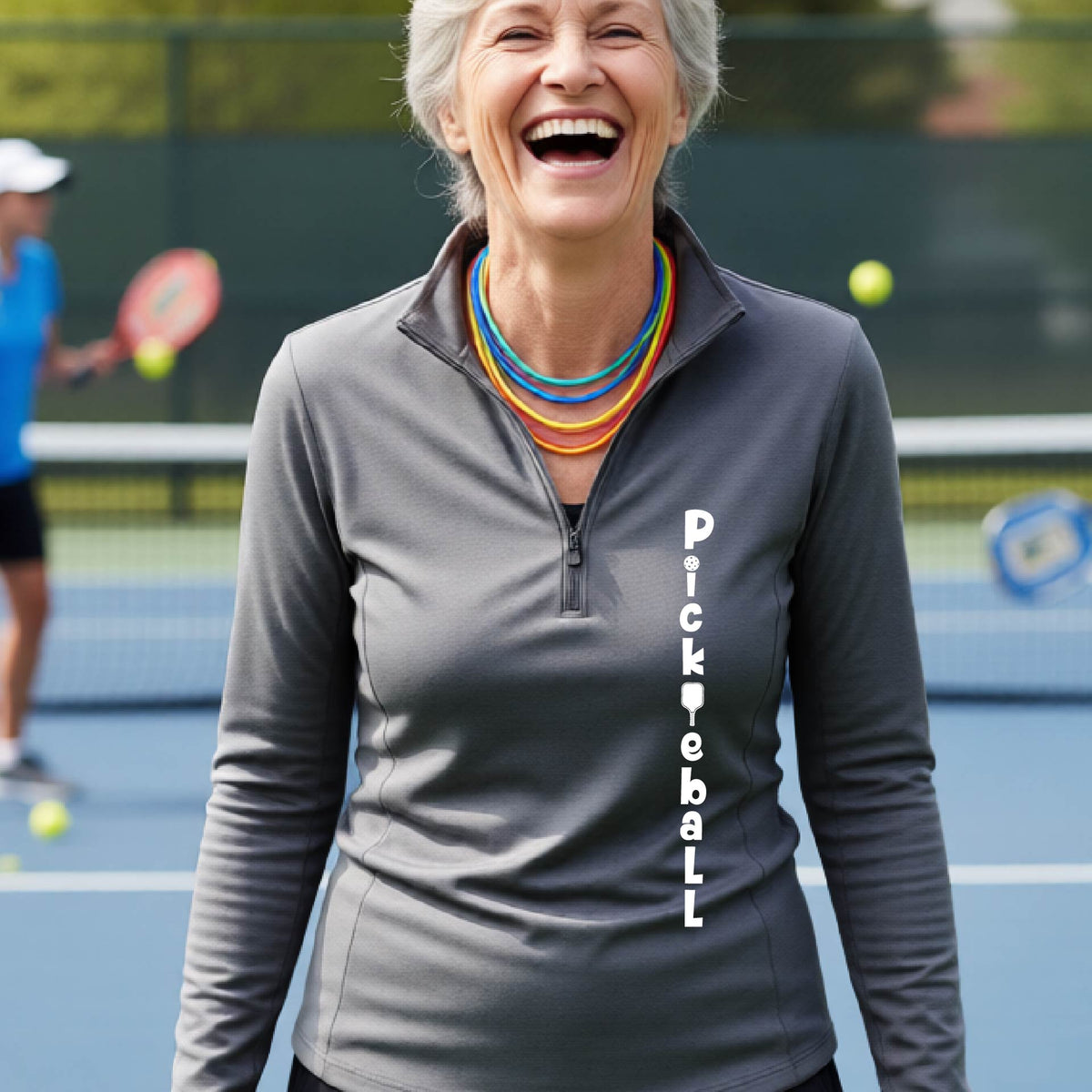 A woman wearing a gray quarter-zip long-sleeve t-shirt with a white graphic text 'PICKLEBALL' on the front with a pickleball paddle replacing the L, standing on a pickleball court.