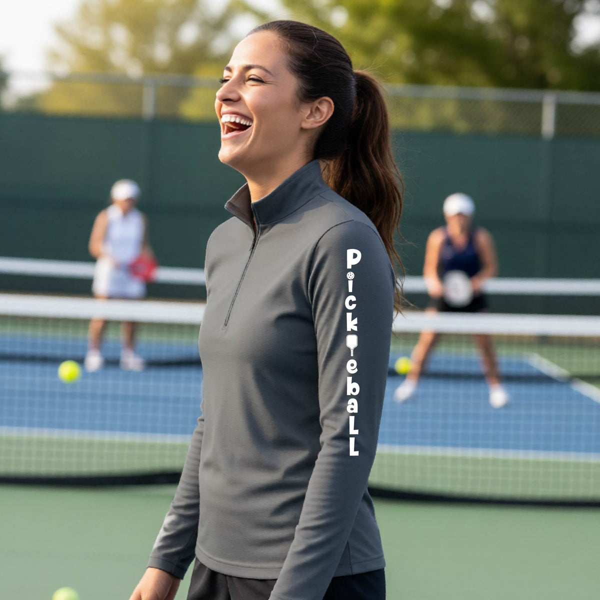A woman wearing a gray quarter-zip long-sleeve t-shirt with a white graphic text 'PICKLEBALL' on the sleeve with a pickleball paddle replacing the L, standing on a pickleball court.