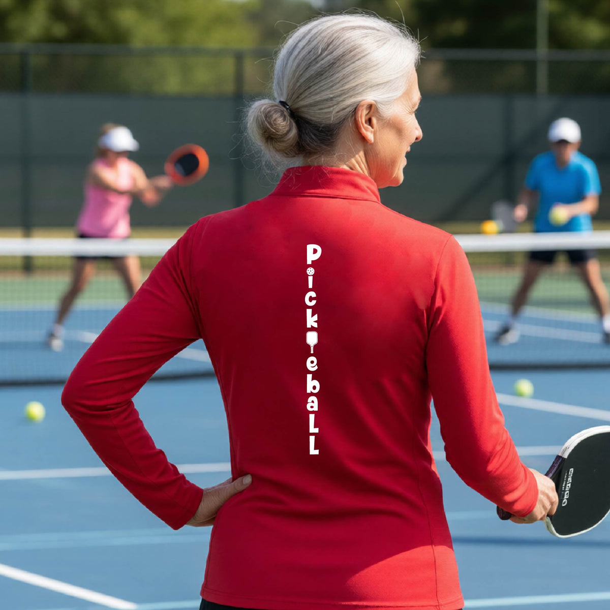 A woman wearing a red quarter-zip long-sleeve t-shirt with a white graphic text 'PICKLEBALL' on the back with a pickleball paddle replacing the L, standing on a pickleball court.