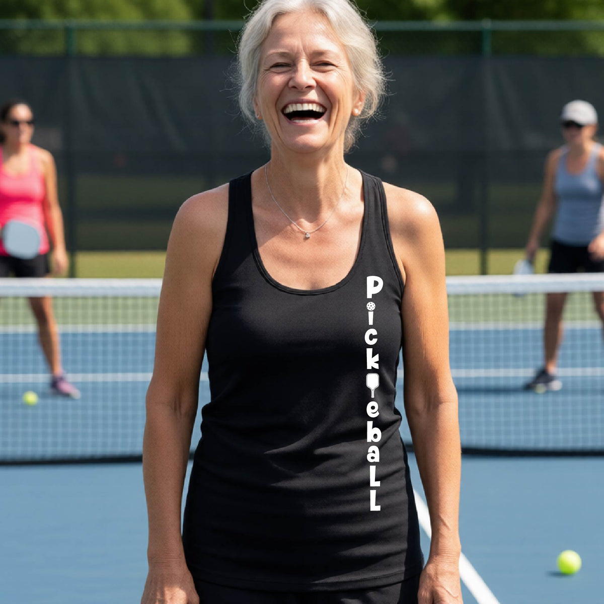 A woman wearing a black racerback tank top with a white graphic text 'PICKLEBALL' on the front with a pickleball paddle replacing the L, standing on a pickleball court.