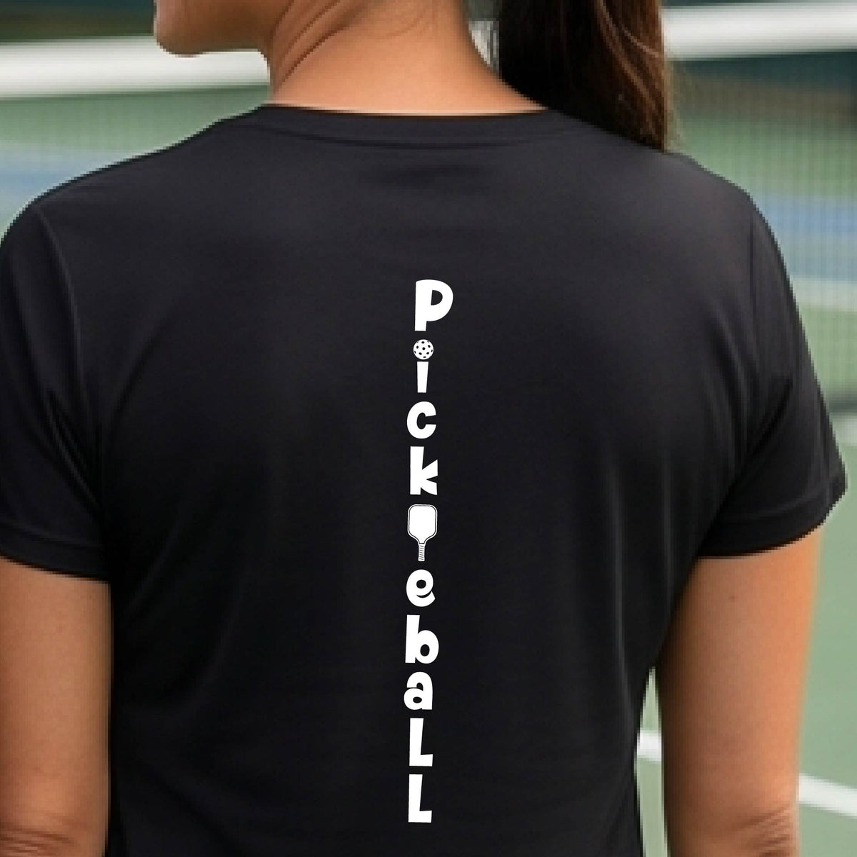 A woman wearing a black short-sleeve t-shirt with a white graphic text 'PICKLEBALL' on the back with a pickleball paddle replacing the L, standing on a pickleball court.