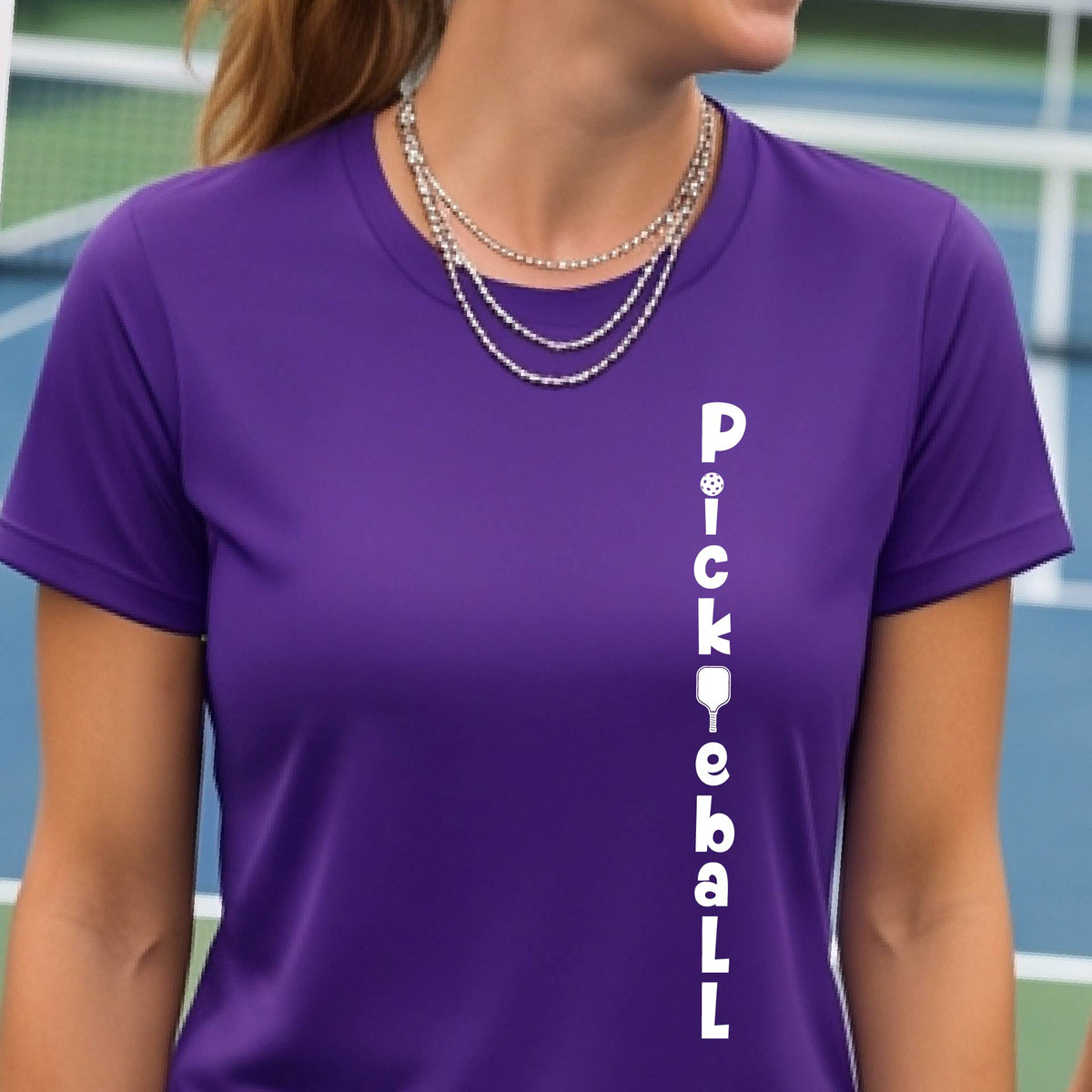 A woman wearing a purple short-sleeve t-shirt with a white graphic text 'PICKLEBALL' on the front with a pickleball paddle replacing the L, standing on a pickleball court.