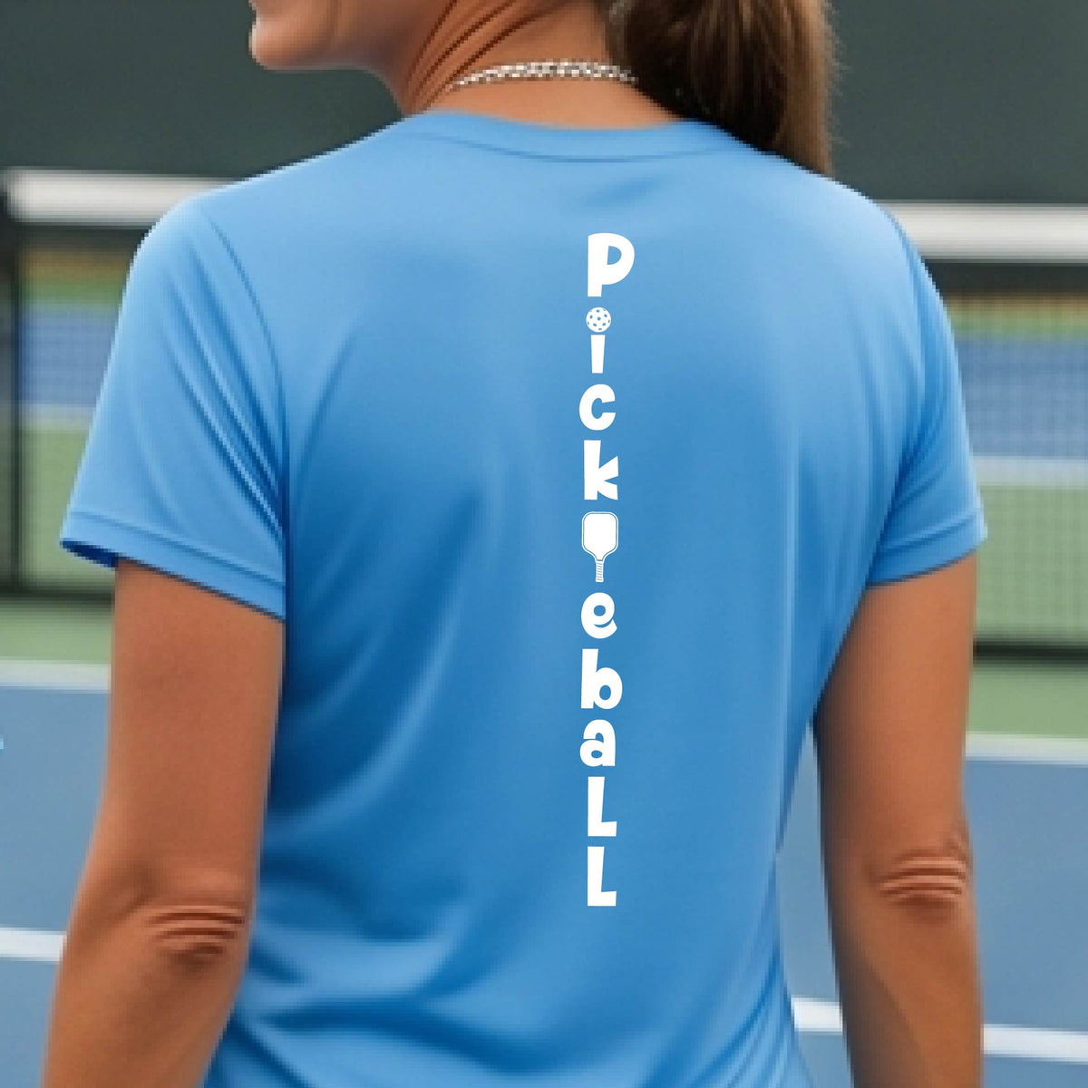 A woman wearing a blue short-sleeve t-shirt with a white graphic text 'PICKLEBALL' on the back with a pickleball paddle replacing the L, standing on a pickleball court.