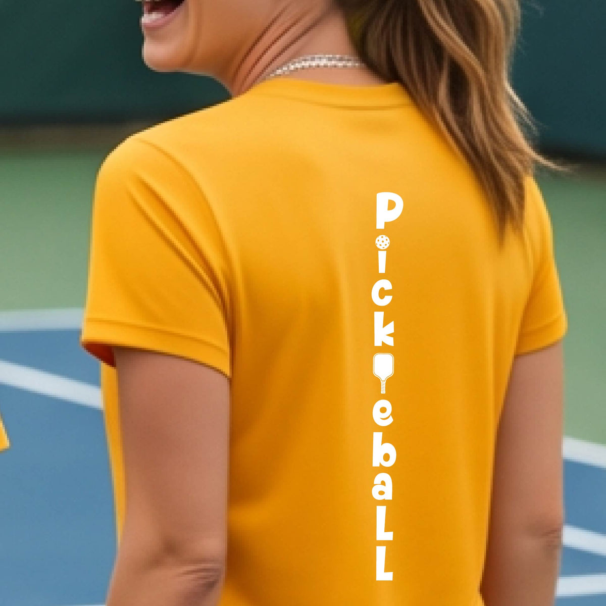 A woman wearing a gold short-sleeve t-shirt with a white graphic text 'PICKLEBALL' on the back with a pickleball paddle replacing the L, standing on a pickleball court.
