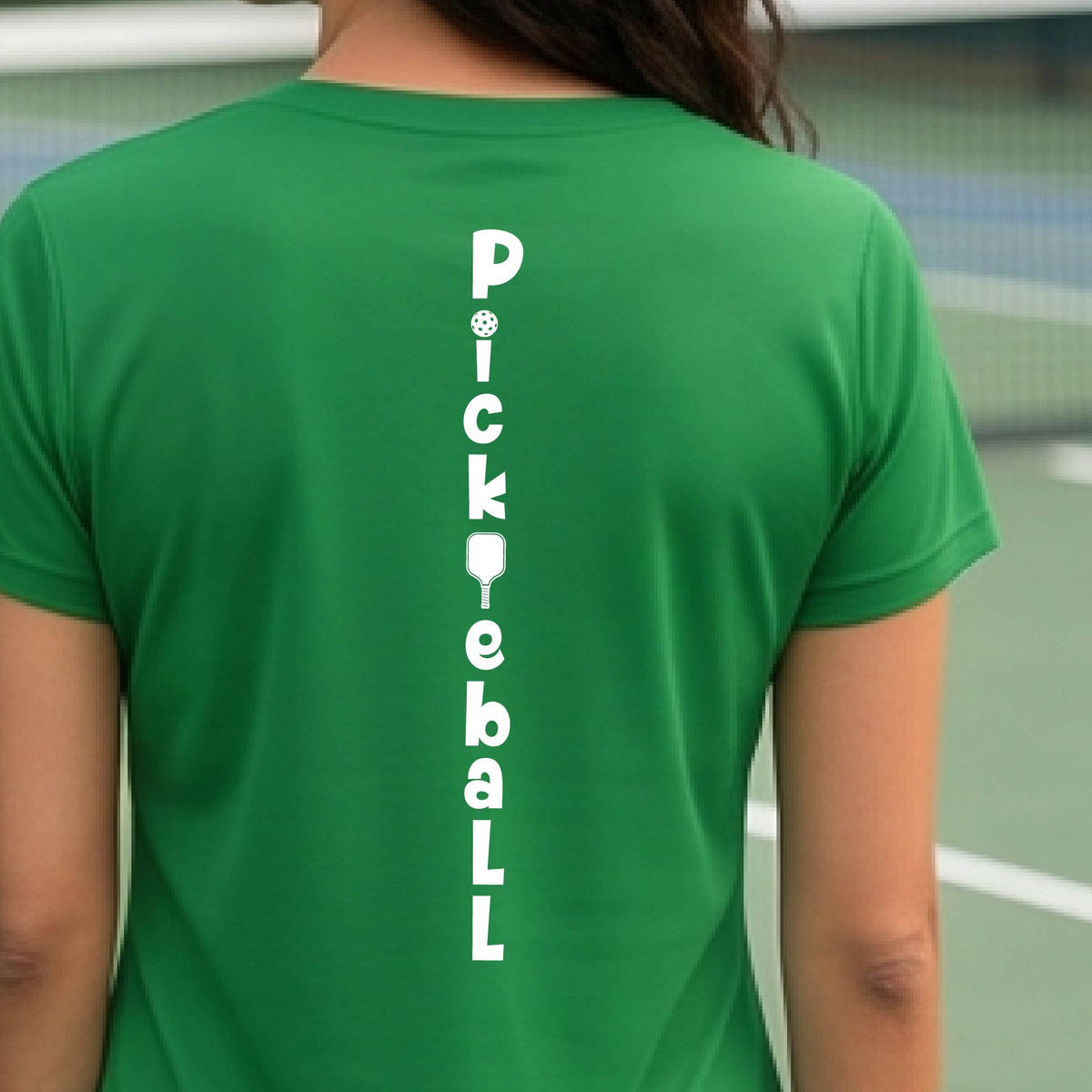A woman wearing a green short-sleeve t-shirt with a white graphic text 'PICKLEBALL' on the back with a pickleball paddle replacing the L, standing on a pickleball court.