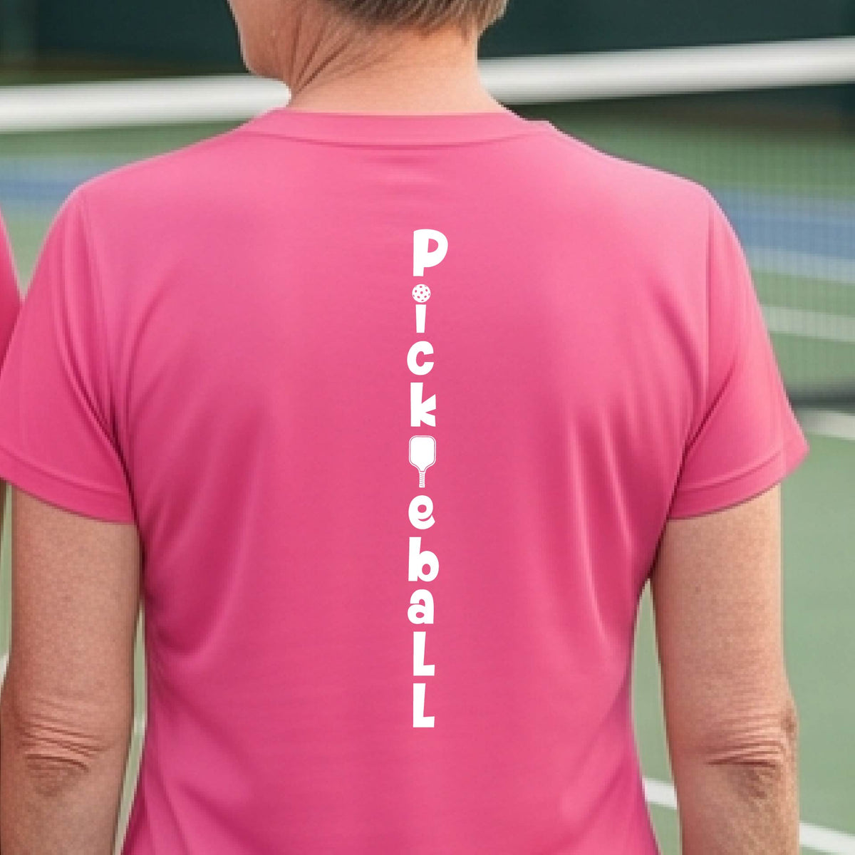 A woman wearing a pink short-sleeve t-shirt with a white graphic text 'PICKLEBALL' on the back with a pickleball paddle replacing the L, standing on a pickleball court.