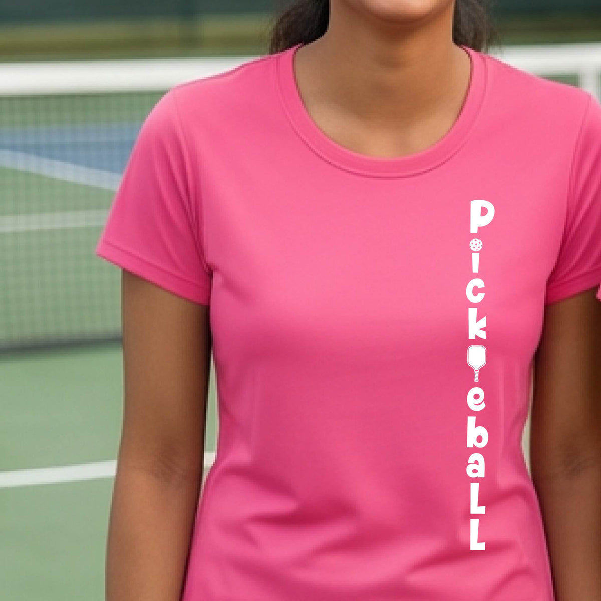 A woman wearing a pink short-sleeve t-shirt with a white graphic text 'PICKLEBALL' on the front with a pickleball paddle replacing the L, standing on a pickleball court.