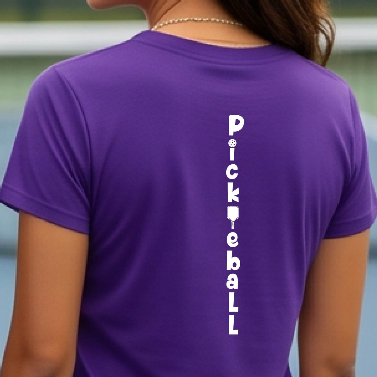A woman wearing a purple short-sleeve t-shirt with a white graphic text 'PICKLEBALL' on the back with a pickleball paddle replacing the L, standing on a pickleball court.
