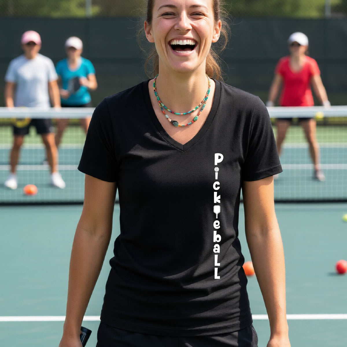 A woman wearing a black short-sleeve v-neck t-shirt with a white graphic text 'PICKLEBALL' on the front with a pickleball paddle replacing the L, standing on a pickleball court.