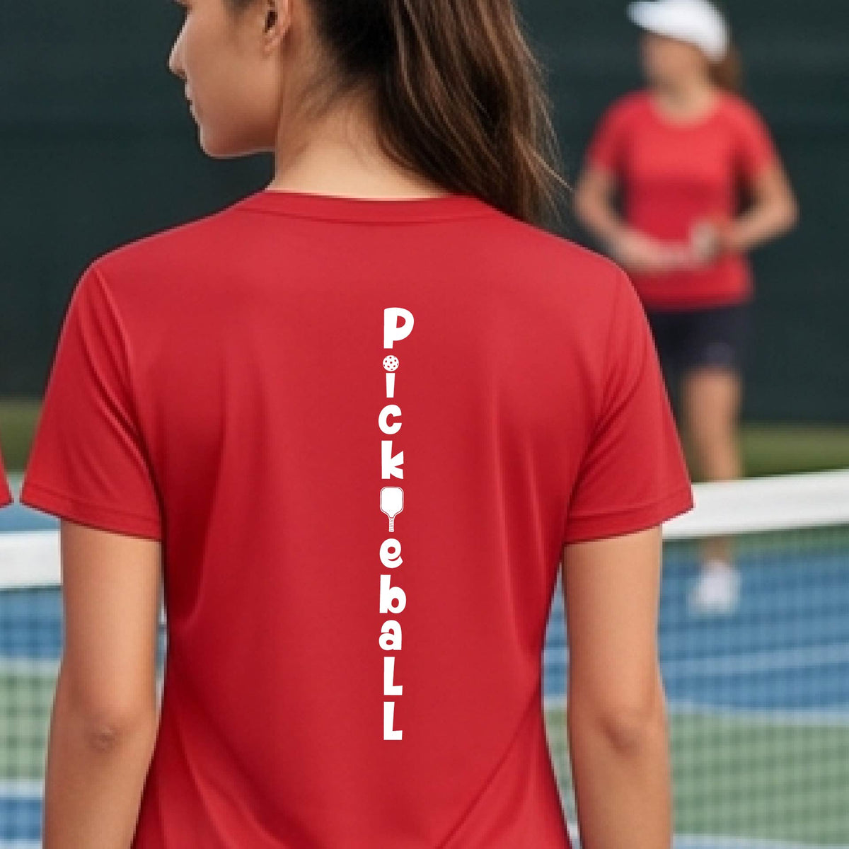 A woman wearing a red short-sleeve v-neck t-shirt with a white graphic text 'PICKLEBALL' on the back with a pickleball paddle replacing the L, standing on a pickleball court.