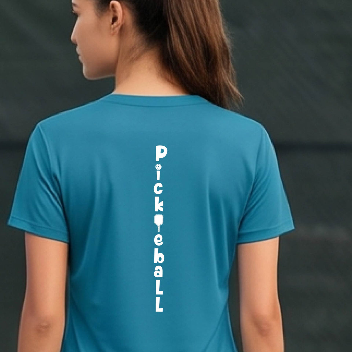 A woman wearing a teal short-sleeve v-neck t-shirt with a white graphic text 'PICKLEBALL' on the back with a pickleball paddle replacing the L, standing on a pickleball court.