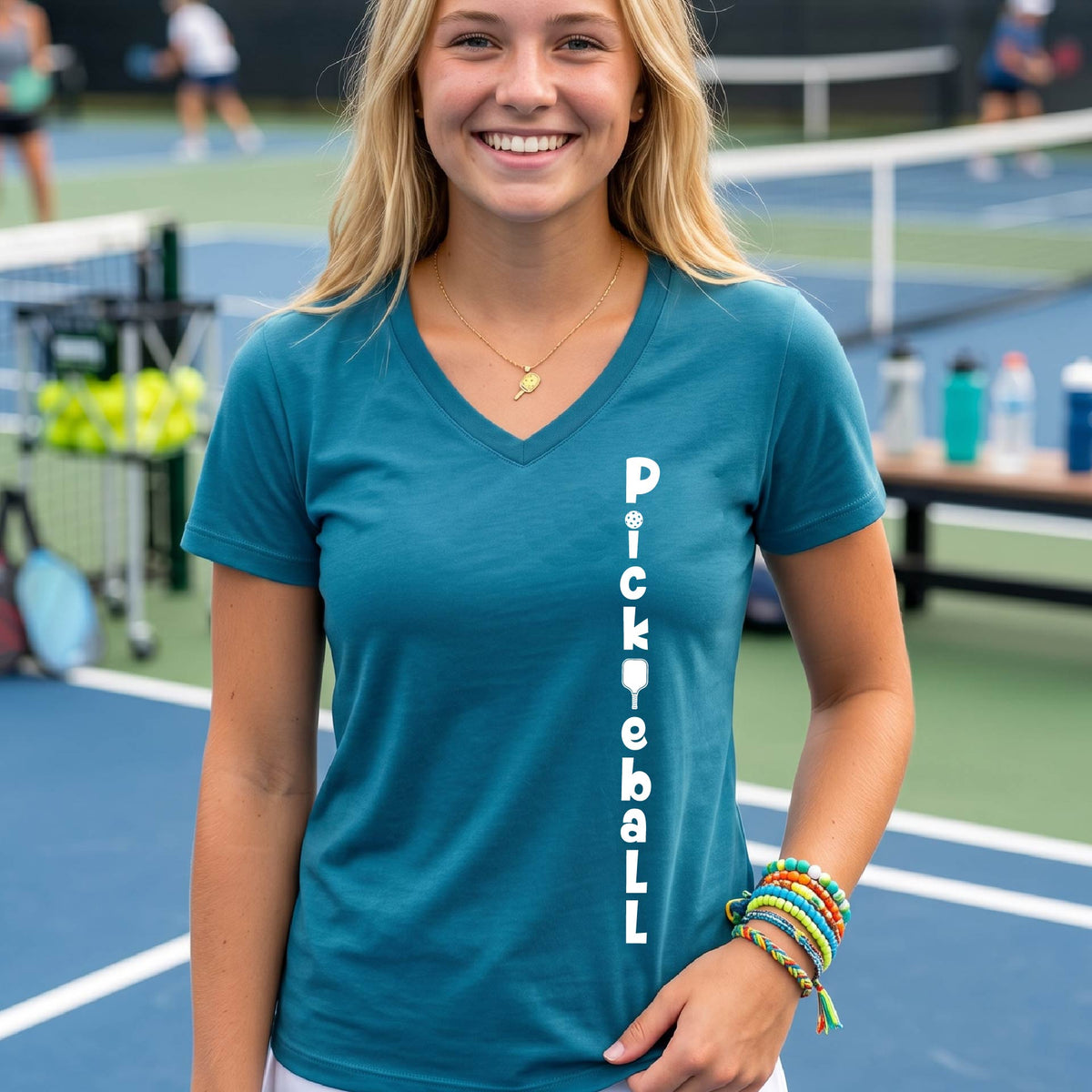 A woman wearing a teal short-sleeve v-neck t-shirt with a white graphic text 'PICKLEBALL' on the front with a pickleball paddle replacing the L, standing on a pickleball court.