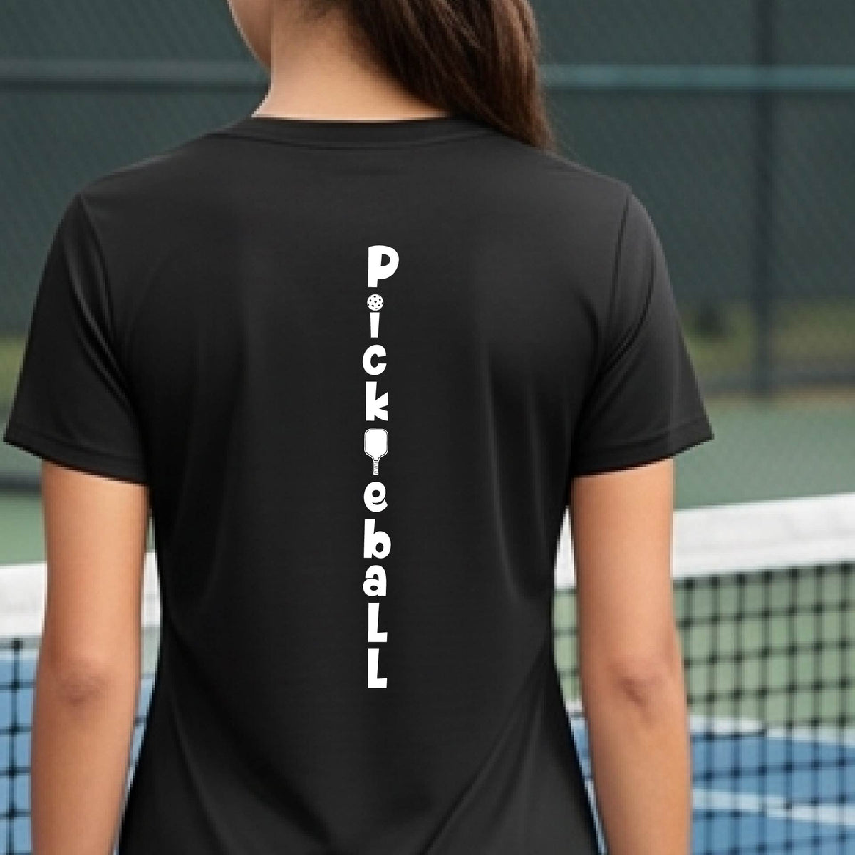 A woman wearing a black short-sleeve v-neck t-shirt with a white graphic text 'PICKLEBALL' on the back with a pickleball paddle replacing the L, standing on a pickleball court.