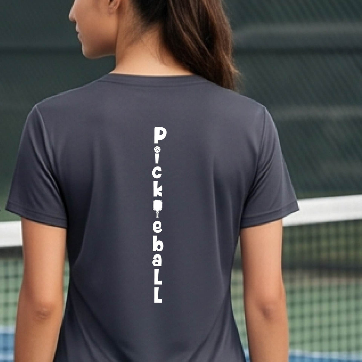 A woman wearing a gray short-sleeve v-neck t-shirt with a white graphic text 'PICKLEBALL' on the back with a pickleball paddle replacing the L, standing on a pickleball court.
