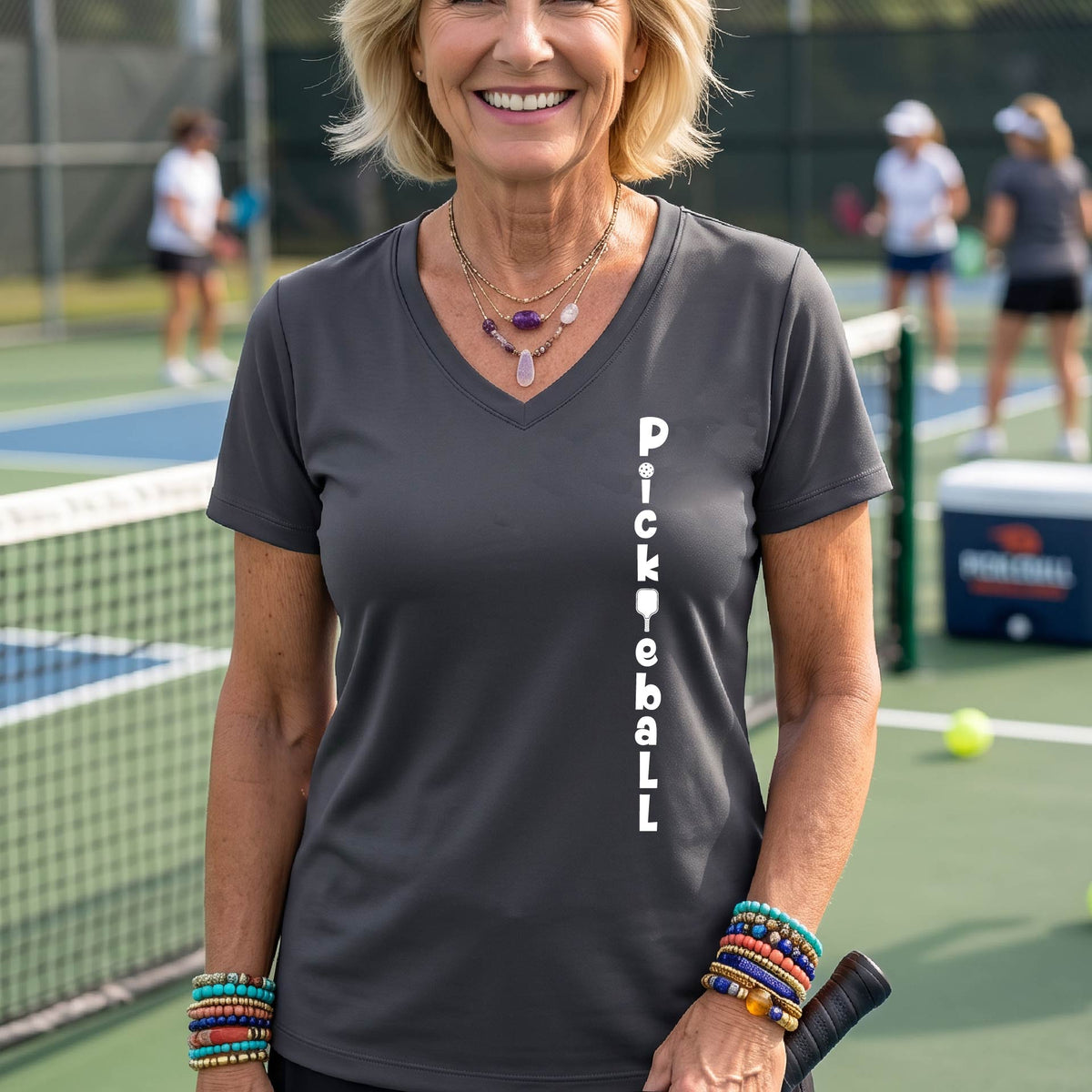 A woman wearing a gray short-sleeve v-neck t-shirt with a white graphic text 'PICKLEBALL' on the front with a pickleball paddle replacing the L, standing on a pickleball court.