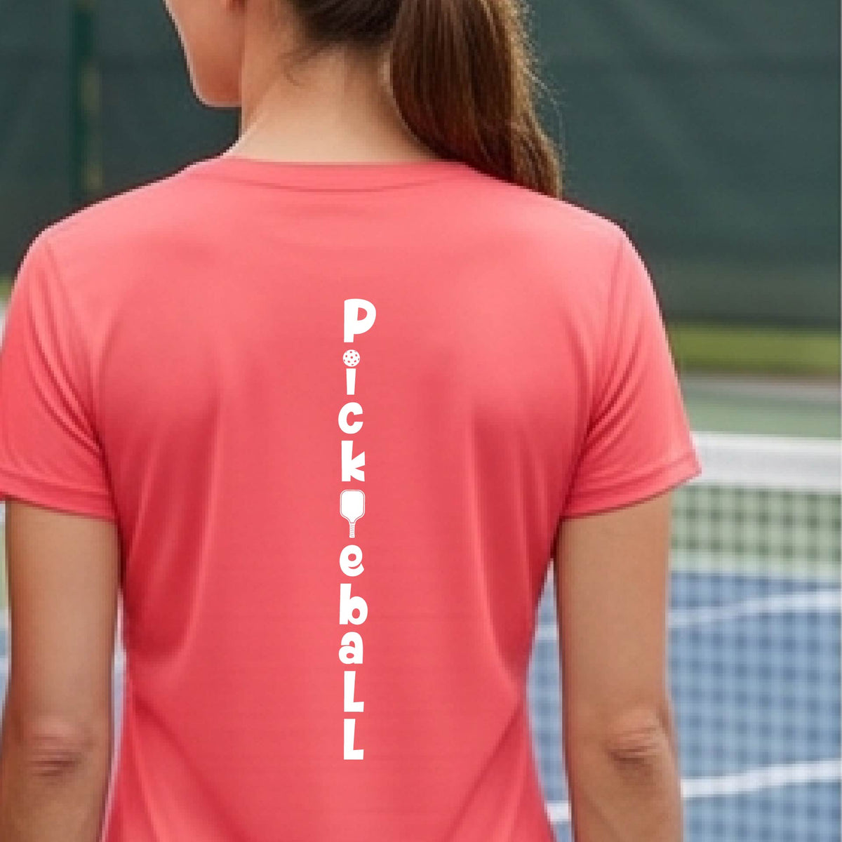A woman wearing a coral pink short-sleeve v-neck t-shirt with a white graphic text 'PICKLEBALL' on the back with a pickleball paddle replacing the L, standing on a pickleball court.