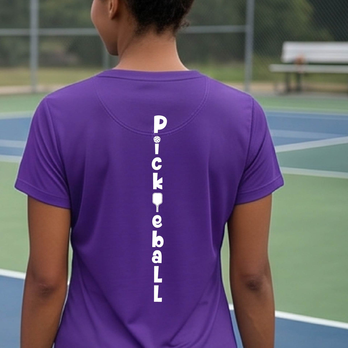 A woman wearing a purple short-sleeve v-neck t-shirt with a white graphic text 'PICKLEBALL' on the back with a pickleball paddle replacing the L, standing on a pickleball court.