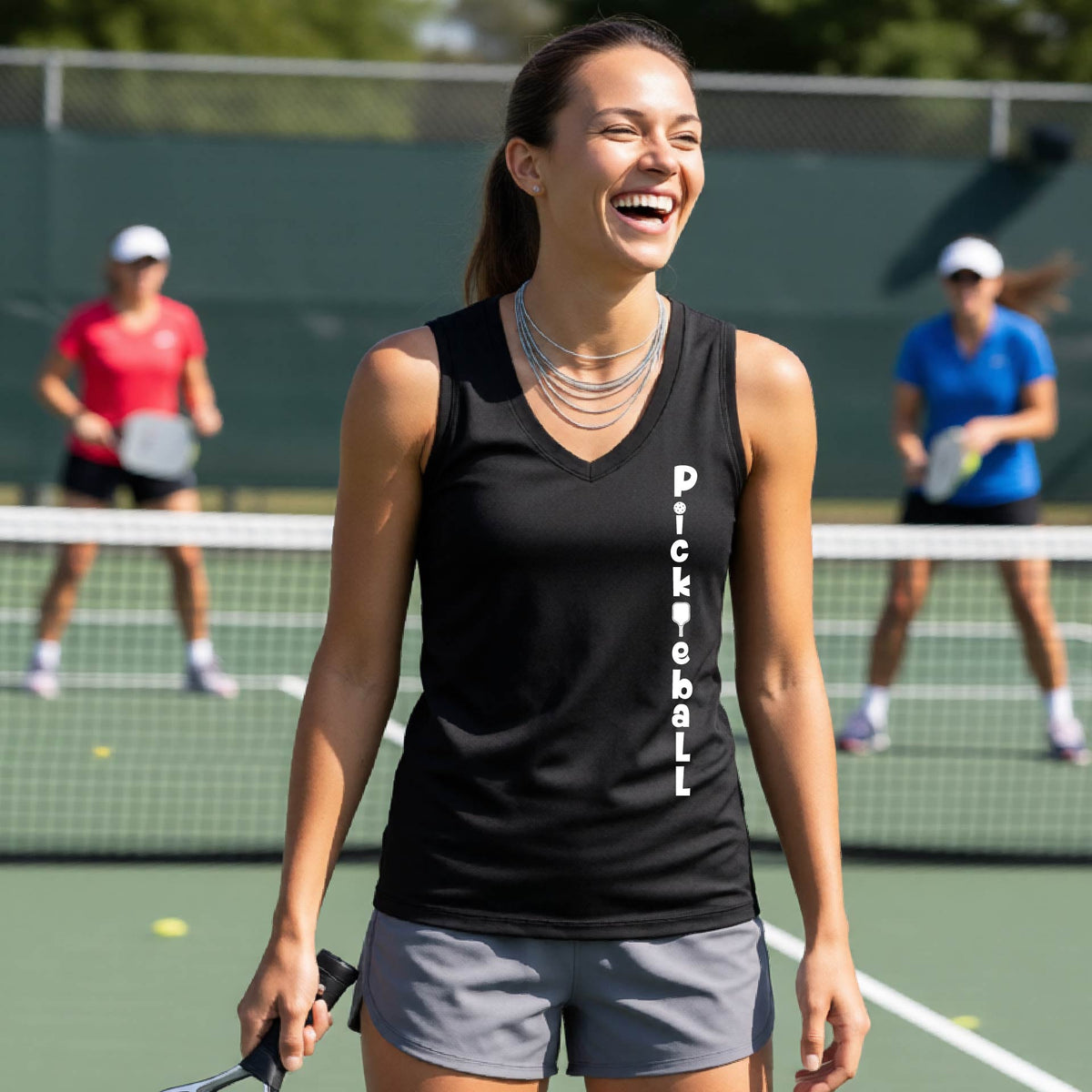 A woman wearing a black sleeveless v-neck t-shirt with a white graphic text 'PICKLEBALL' on the front with a pickleball paddle replacing the L, standing on a pickleball court.