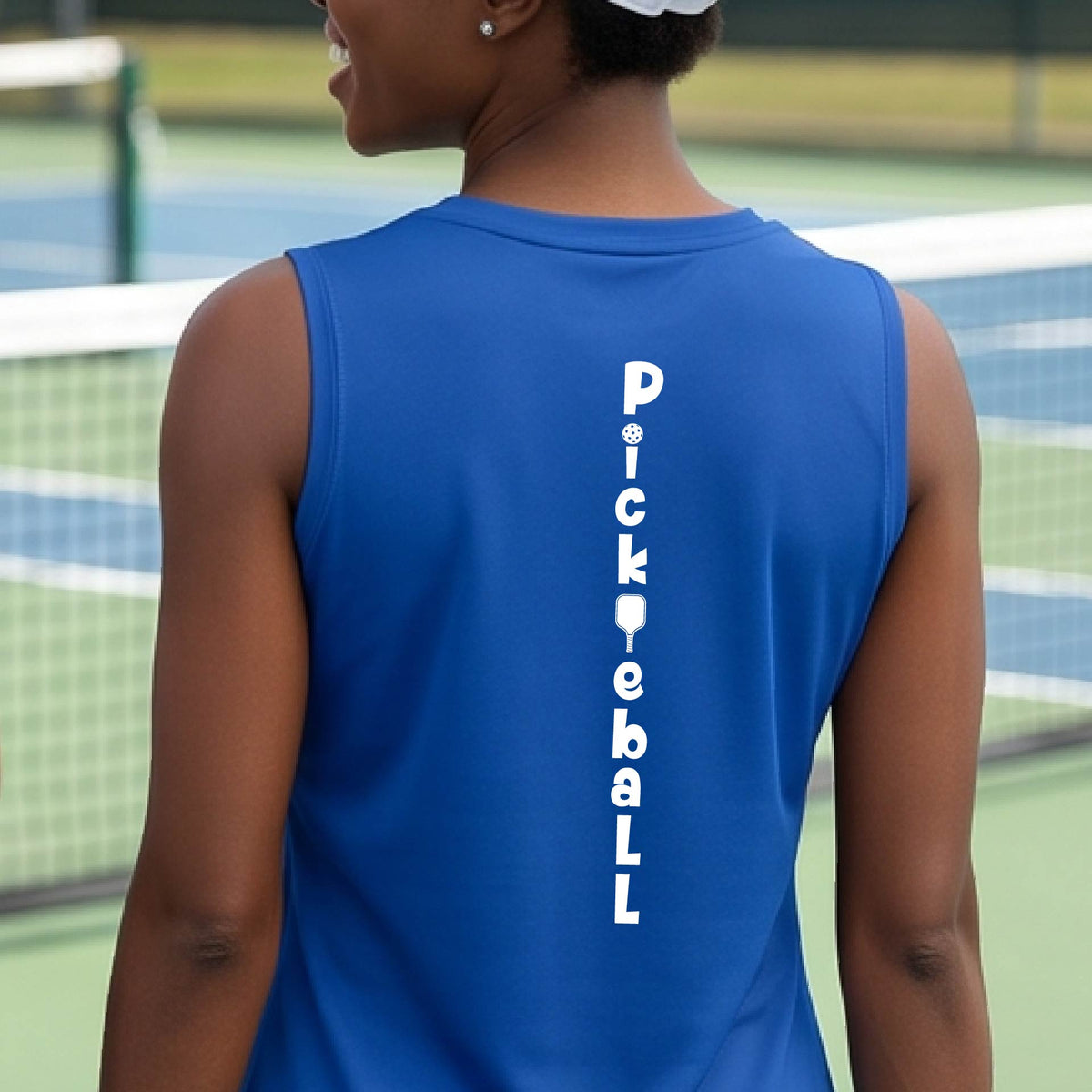 A woman wearing a royal blue sleeveless v-neck t-shirt with a white graphic text 'PICKLEBALL' on the back with a pickleball paddle replacing the L, standing on a pickleball court.