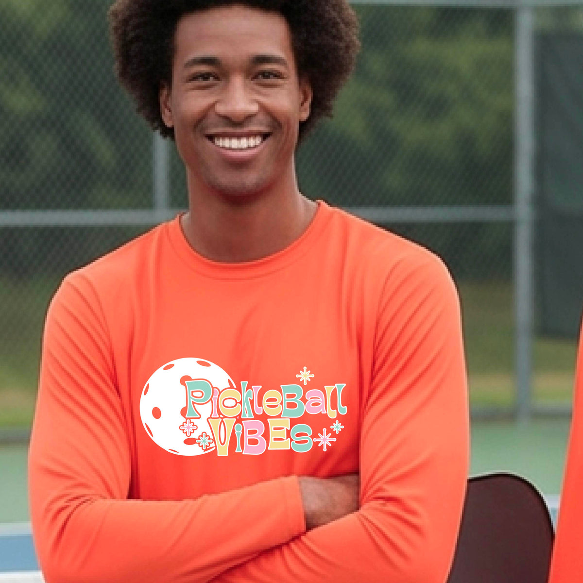 A close-up of a man on a pickleball wearing an orange short-sleeve t-shirt, with a colorful retro style font 'Pickleball Vibes' on top of a white pickleball graphic, on the front of the shirt.