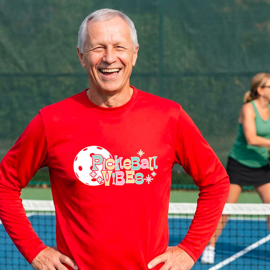 A close-up of a man on a pickleball wearing a red short-sleeve t-shirt, with a colorful retro style font 'Pickleball Vibes' on top of a white pickleball graphic, on the front of the shirt.