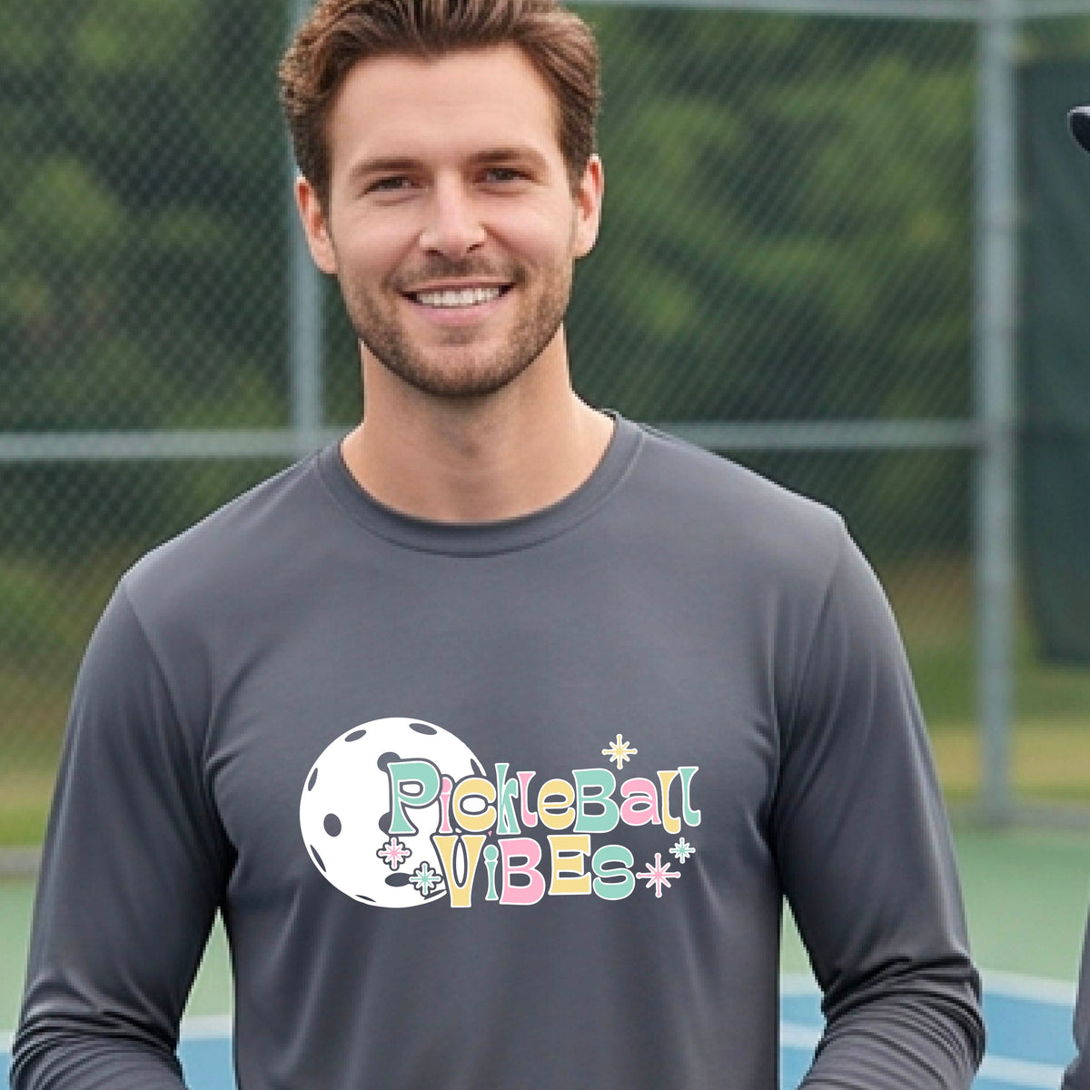 A close-up of a man on a pickleball wearing a gray short-sleeve t-shirt, with a colorful retro style font 'Pickleball Vibes' on top of a white pickleball graphic, on the front of the shirt.