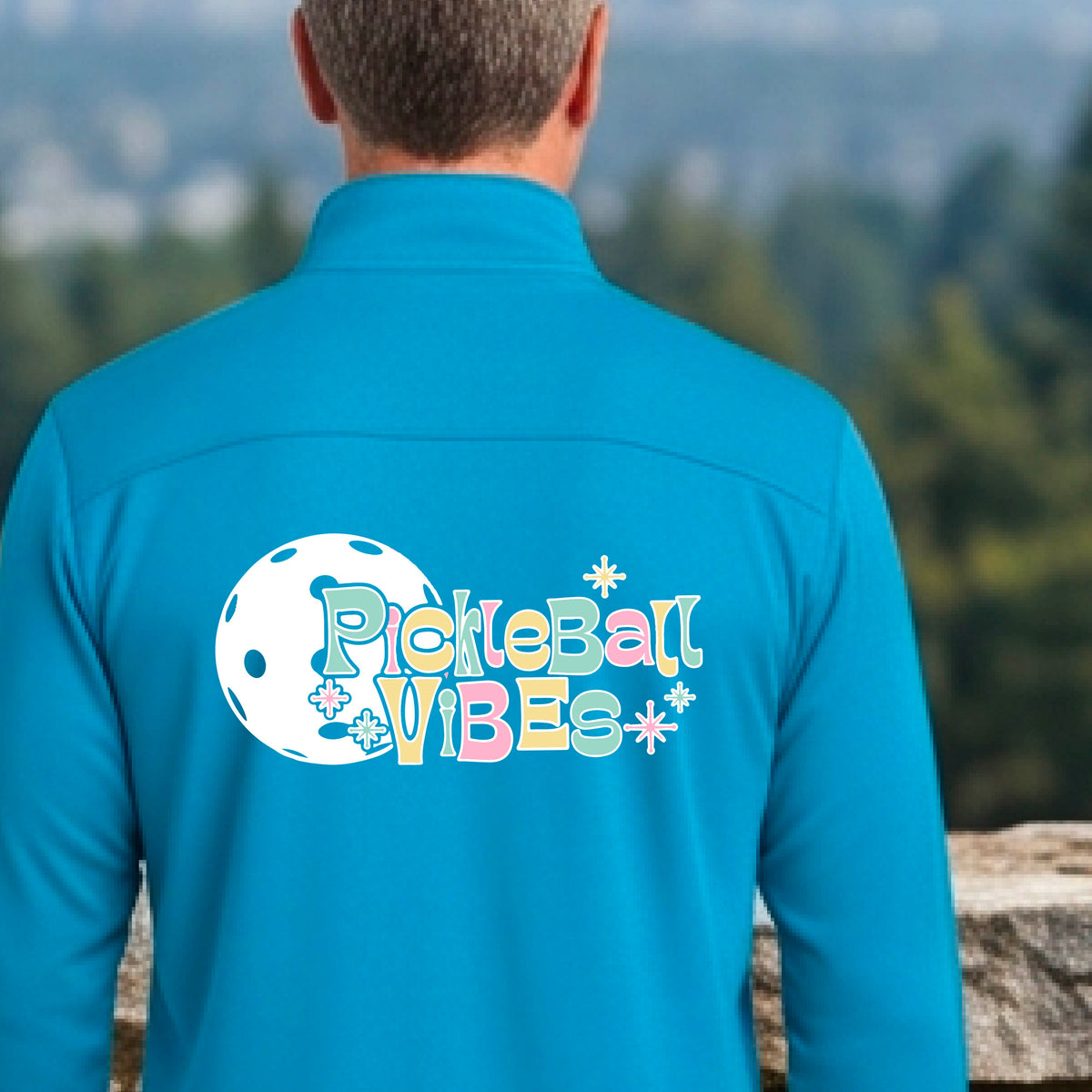 A close-up of a man, facing backwards on a pickleball court wearing a bright blue quarter-zip shirt, with a colorful retro style font 'Pickleball Vibes' on top of a white pickleball graphic, on the back of the shirt.