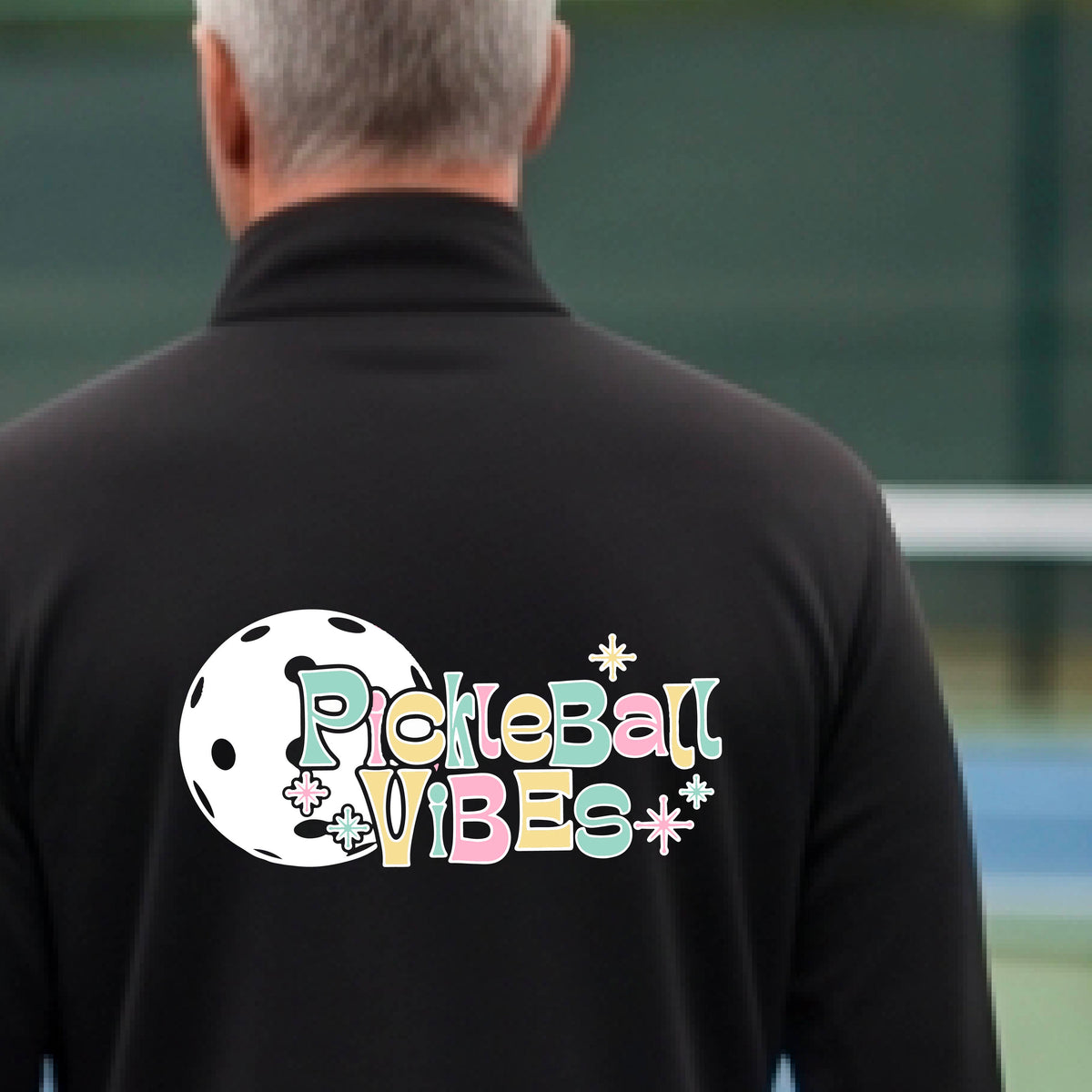 A close-up of a man, facing backwards on a pickleball court wearing a black quarter-zip shirt, with a colorful retro style font 'Pickleball Vibes' on top of a white pickleball graphic, on the back of the shirt.