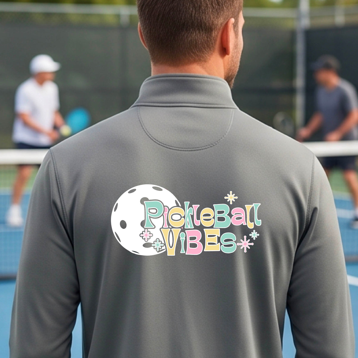 A close-up of a man, facing backwards on a pickleball court wearing a gray quarter-zip shirt, with a colorful retro style font 'Pickleball Vibes' on top of a white pickleball graphic, on the back of the shirt.