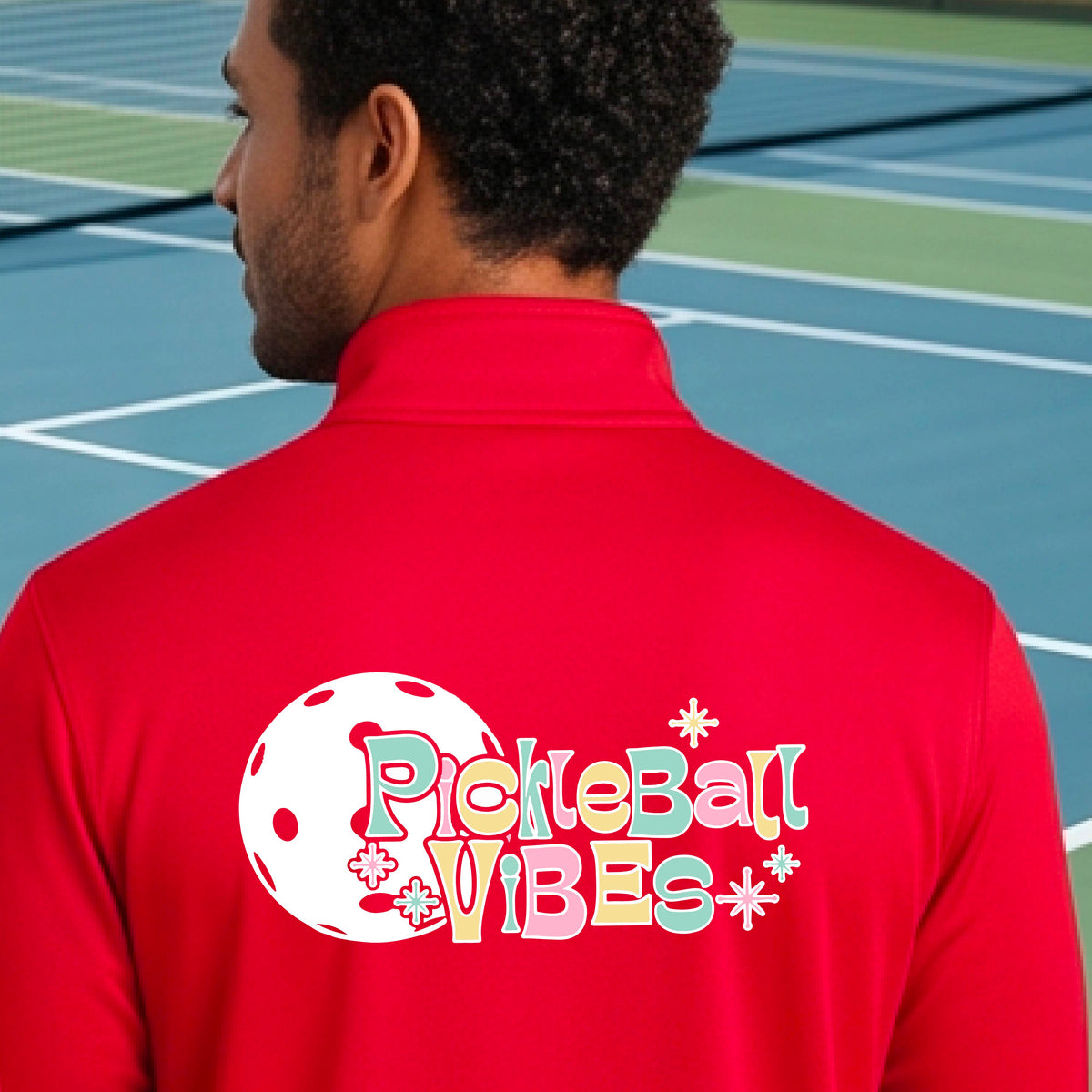 A close-up of a man, facing backwards on a pickleball court wearing a red quarter-zip shirt, with a colorful retro style font 'Pickleball Vibes' on top of a white pickleball graphic, on the back of the shirt.