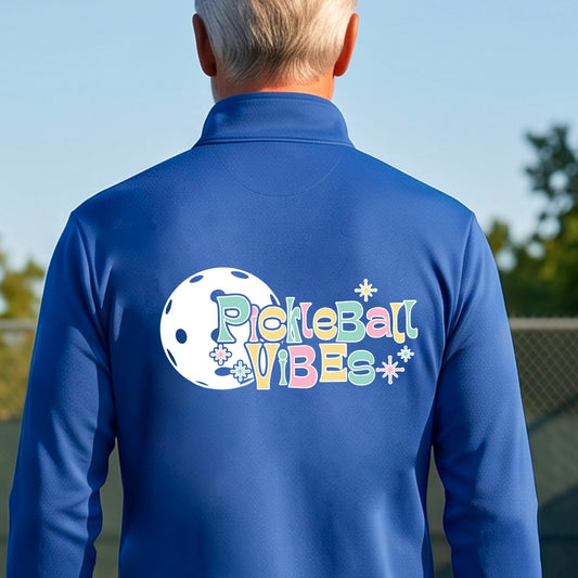 A close-up of a man, facing backwards on a pickleball court wearing a royal blue quarter-zip shirt, with a colorful retro style font 'Pickleball Vibes' on top of a white pickleball graphic, on the back of the shirt.