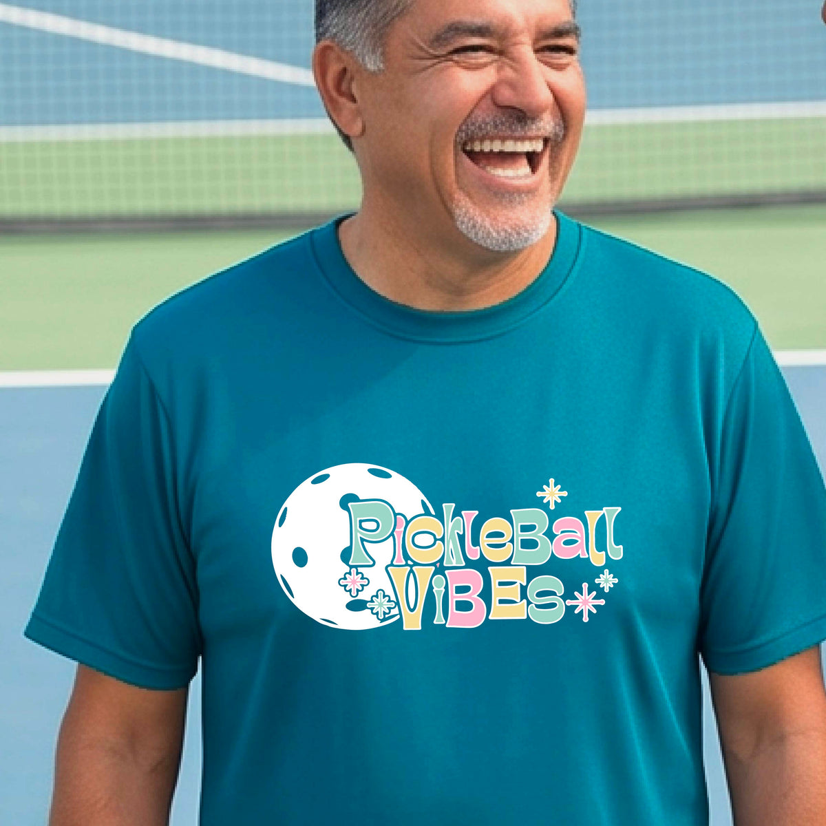 A close-up of a smiling man on a pickleball wearing a teal short-sleeve t-shirt, with a colorful retro style font 'Pickleball Vibes' on top of a white pickleball graphic, on the front of the shirt.