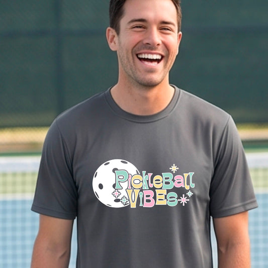 A close-up of a smiling man on a pickleball wearing a gray short-sleeve t-shirt, with a colorful retro style font 'Pickleball Vibes' on top of a white pickleball graphic, on the front of the shirt.