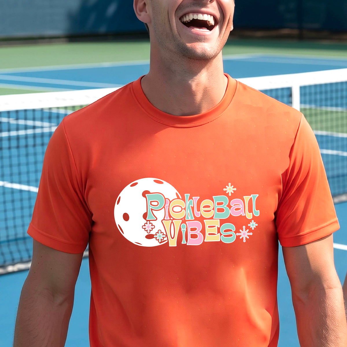 A close-up of a smiling man on a pickleball wearing an orange short-sleeve t-shirt, with a colorful retro style font 'Pickleball Vibes' on top of a white pickleball graphic, on the front of the shirt.