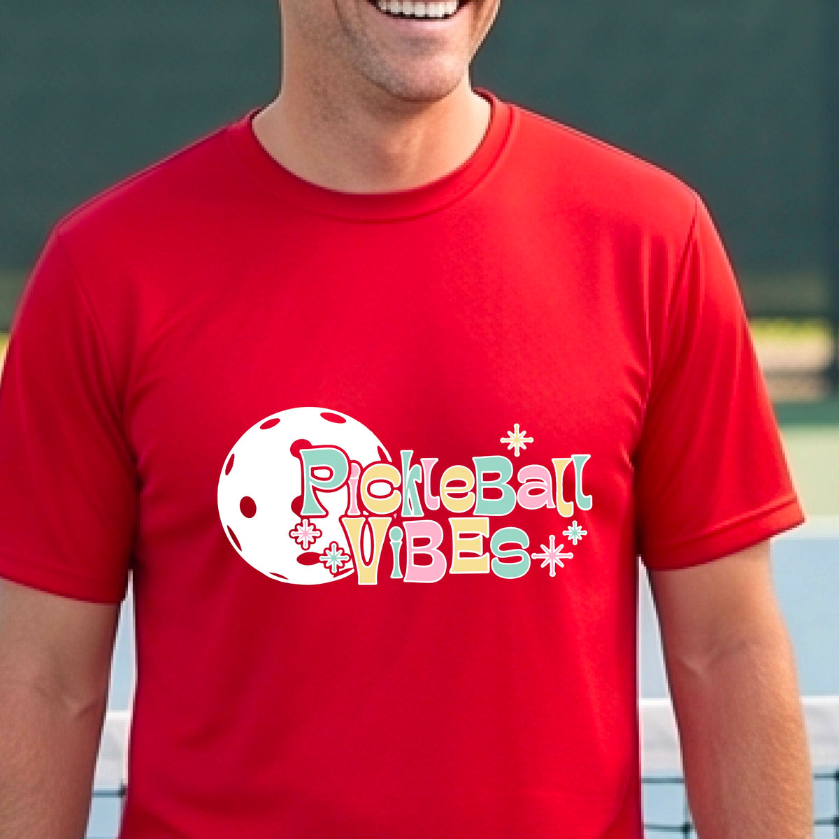 A close-up of a smiling man on a pickleball wearing a red short-sleeve t-shirt, with a colorful retro style font 'Pickleball Vibes' on top of a white pickleball graphic, on the front of the shirt.