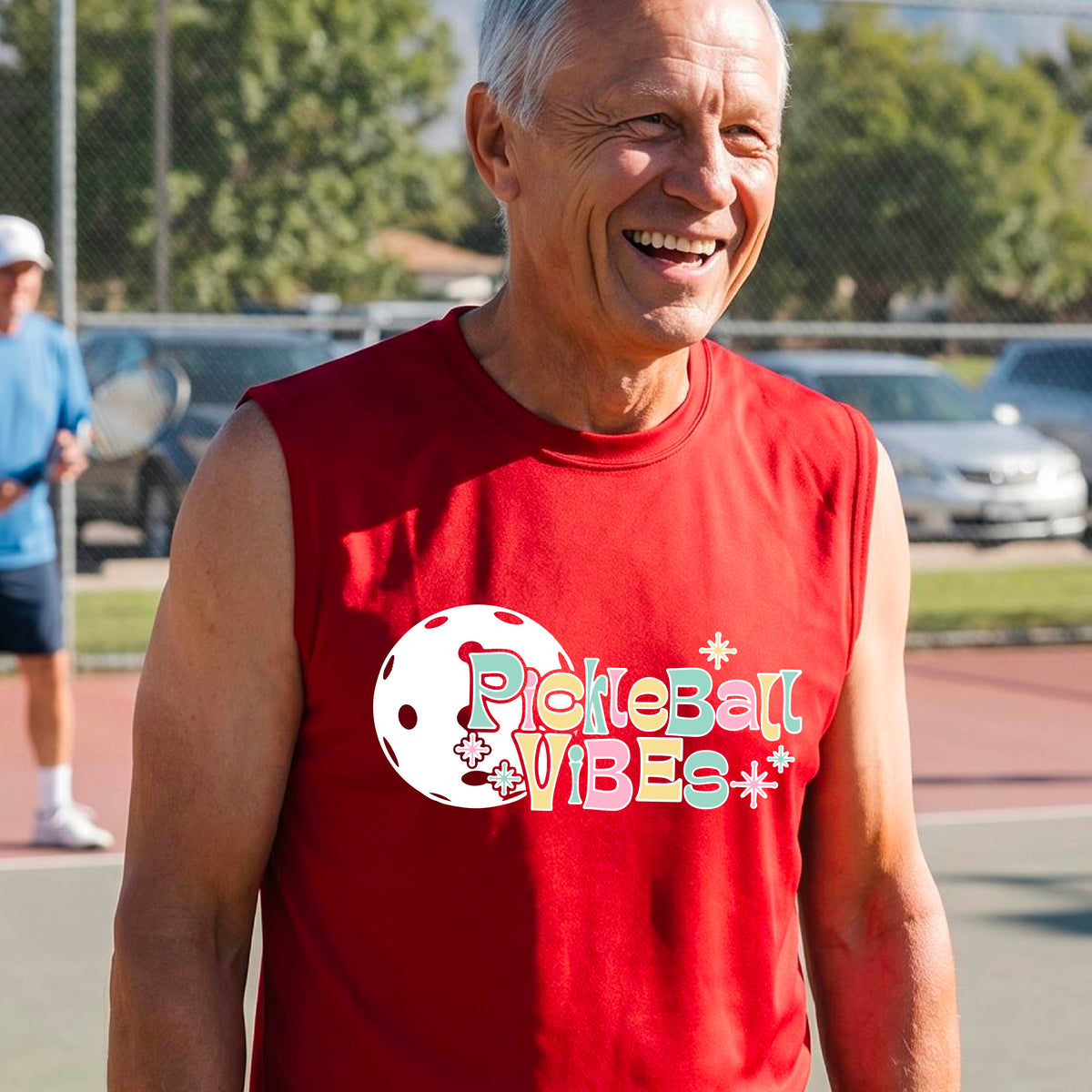 A close-up of a man on a pickleball wearing a red sleeveless t-shirt, with a colorful retro style font 'Pickleball Vibes' on top of a white pickleball graphic, on the front of the shirt.