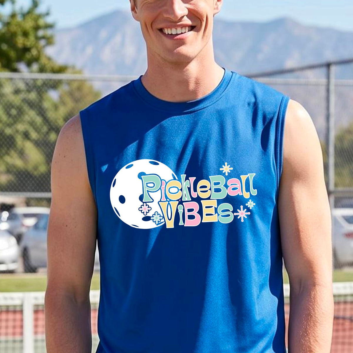 A close-up of a man on a pickleball wearing a royal blue sleeveless t-shirt, with a colorful retro style font 'Pickleball Vibes' on top of a white pickleball graphic, on the front of the shirt.