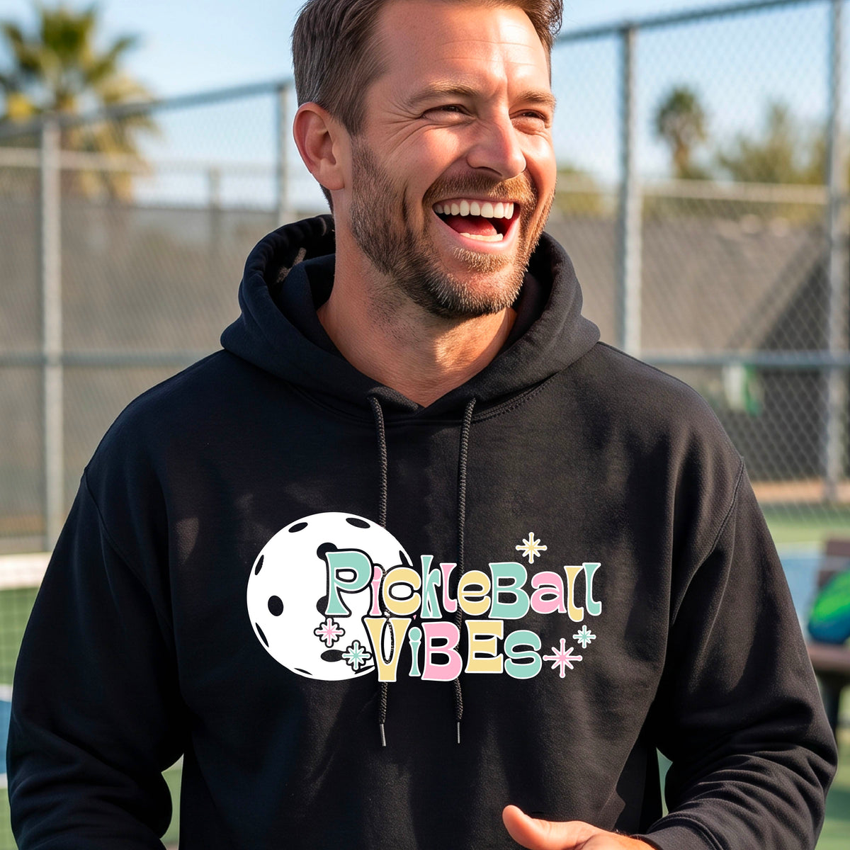 A close-up of a man on a pickleball court wearing a black hoodie pullover, with a colorful retro style font 'Pickleball Vibes' on top of a white pickleball graphic, on the front of the hoodie. 