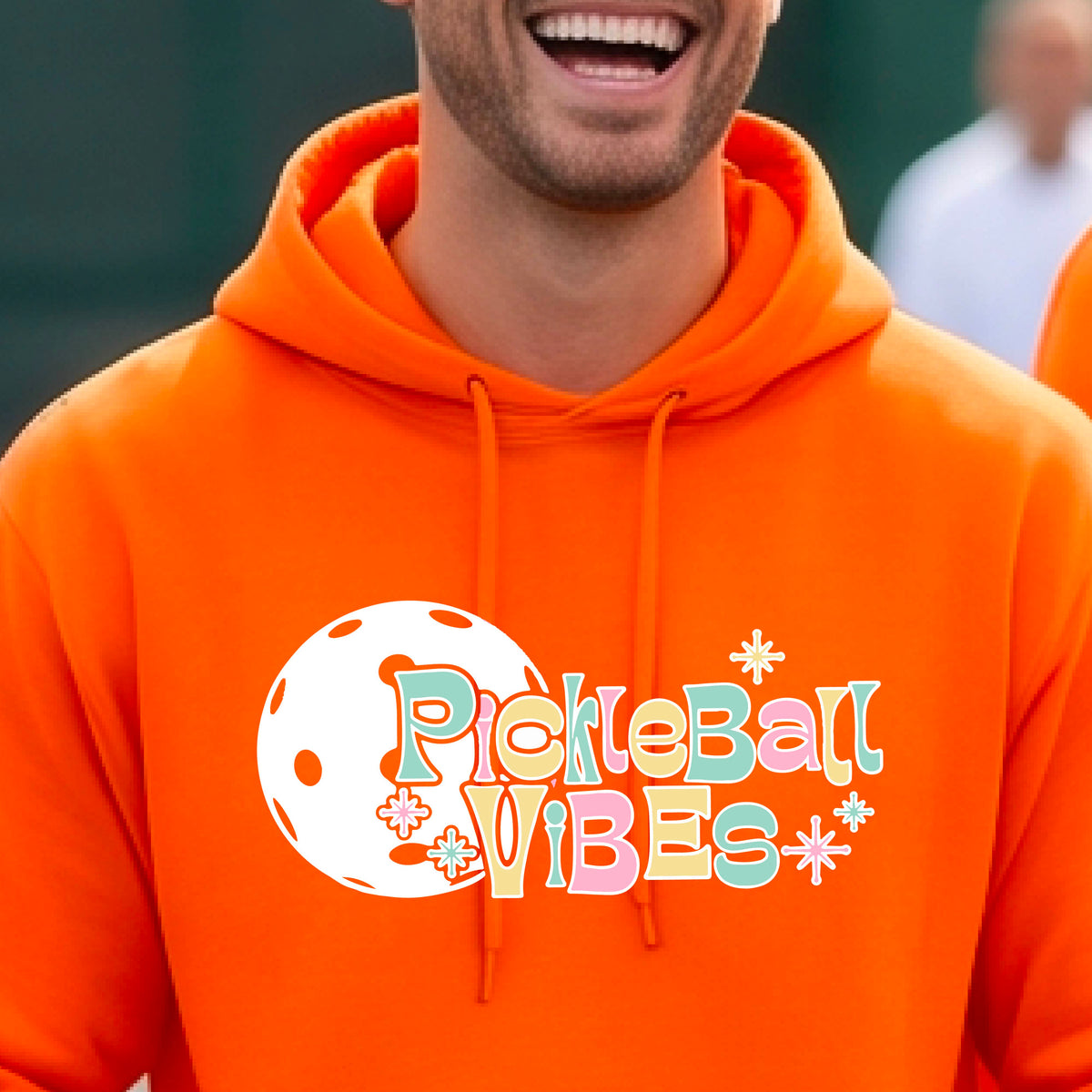 A close-up of a man on a pickleball court wearing an orange hoodie pullover, with a colorful retro style font 'Pickleball Vibes' on top of a white pickleball graphic, on the front of the hoodie. 