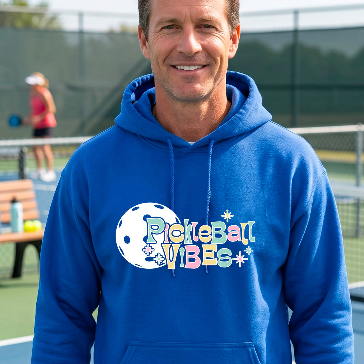 A close-up of a man on a pickleball court wearing a royal blue hoodie pullover, with a colorful retro style font 'Pickleball Vibes' on top of a white pickleball graphic, on the front of the hoodie. 