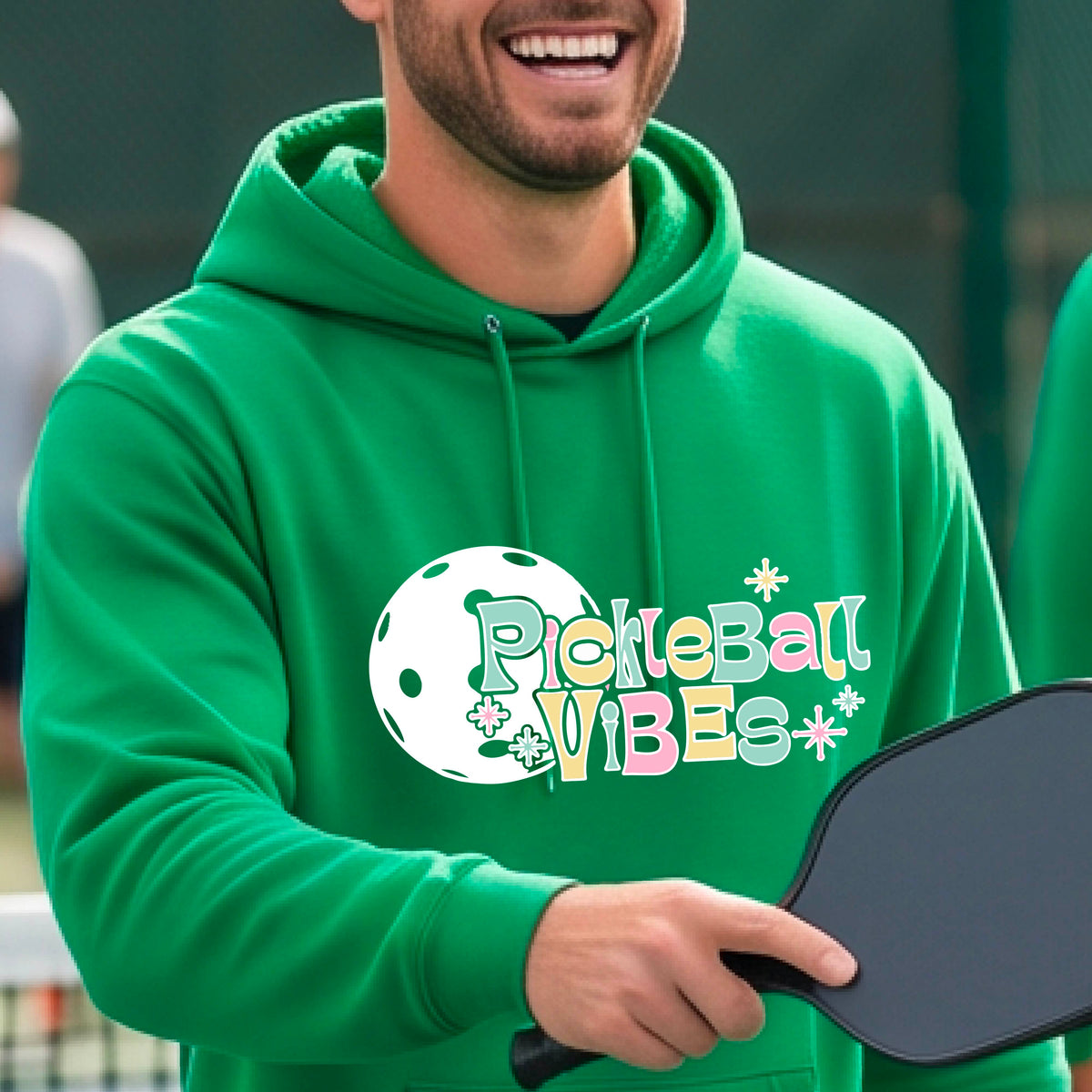 A close-up of a man on a pickleball court wearing a green hoodie pullover, with a colorful retro style font 'Pickleball Vibes' on top of a white pickleball graphic, on the front of the hoodie. 