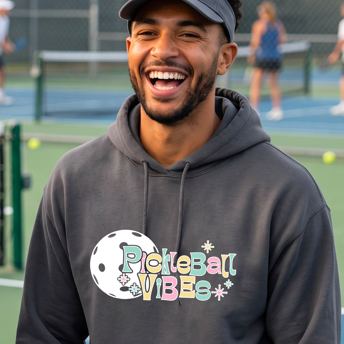 A close-up of a man on a pickleball court wearing a dark gray hoodie pullover, with a colorful retro style font 'Pickleball Vibes' on top of a white pickleball graphic, on the front of the hoodie. 