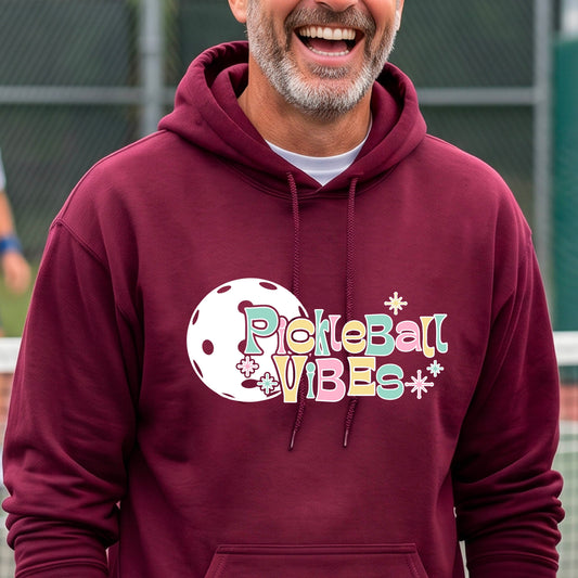 A close-up of a man on a pickleball court wearing a maroon hoodie pullover, with a colorful retro style font 'Pickleball Vibes' on top of a white pickleball graphic, on the front of the hoodie. 