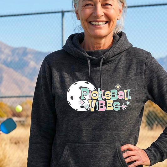 A close-up of a woman on a pickleball court wearing a black cowl-neck hoodie pullover, with a colorful retro style font 'Pickleball Vibes' on top of a white pickleball graphic, on the front of the hoodie. 