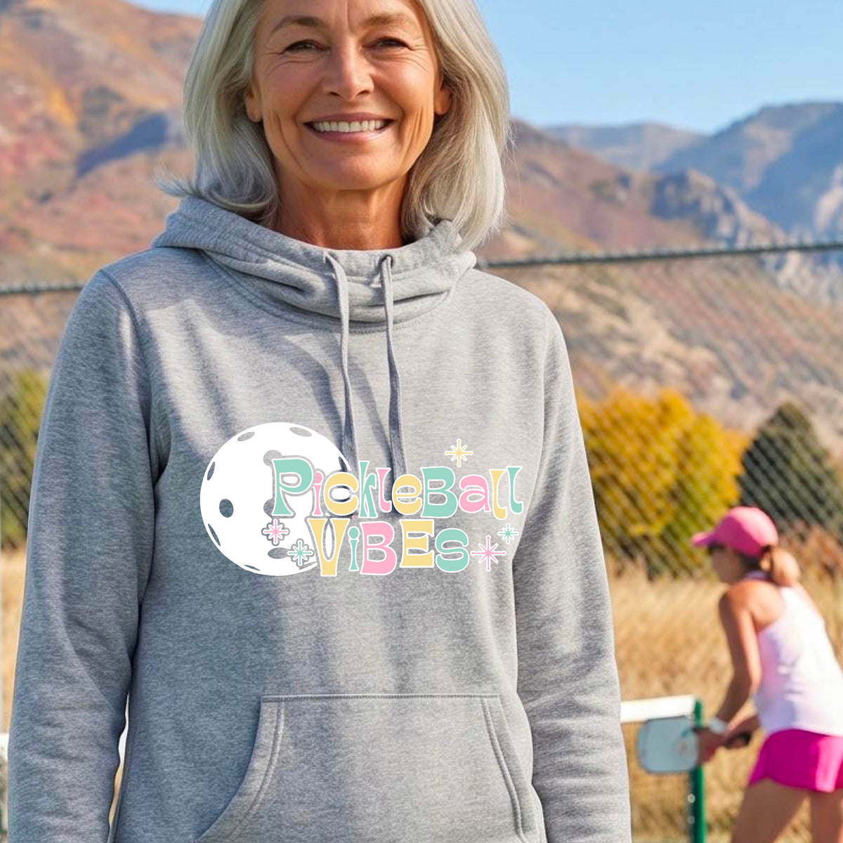 A close-up of a woman on a pickleball court wearing a light gray cowl-neck hoodie pullover, with a colorful retro style font 'Pickleball Vibes' on top of a white pickleball graphic, on the front of the hoodie. 
