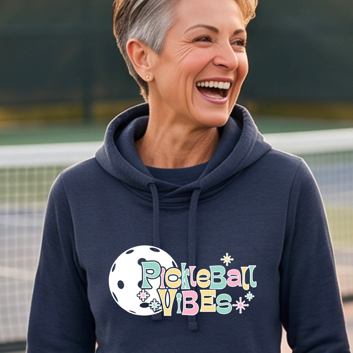 A close-up of a woman on a pickleball court wearing a navy blue cowl-neck hoodie pullover, with a colorful retro style font 'Pickleball Vibes' on top of a white pickleball graphic, on the front of the hoodie. 