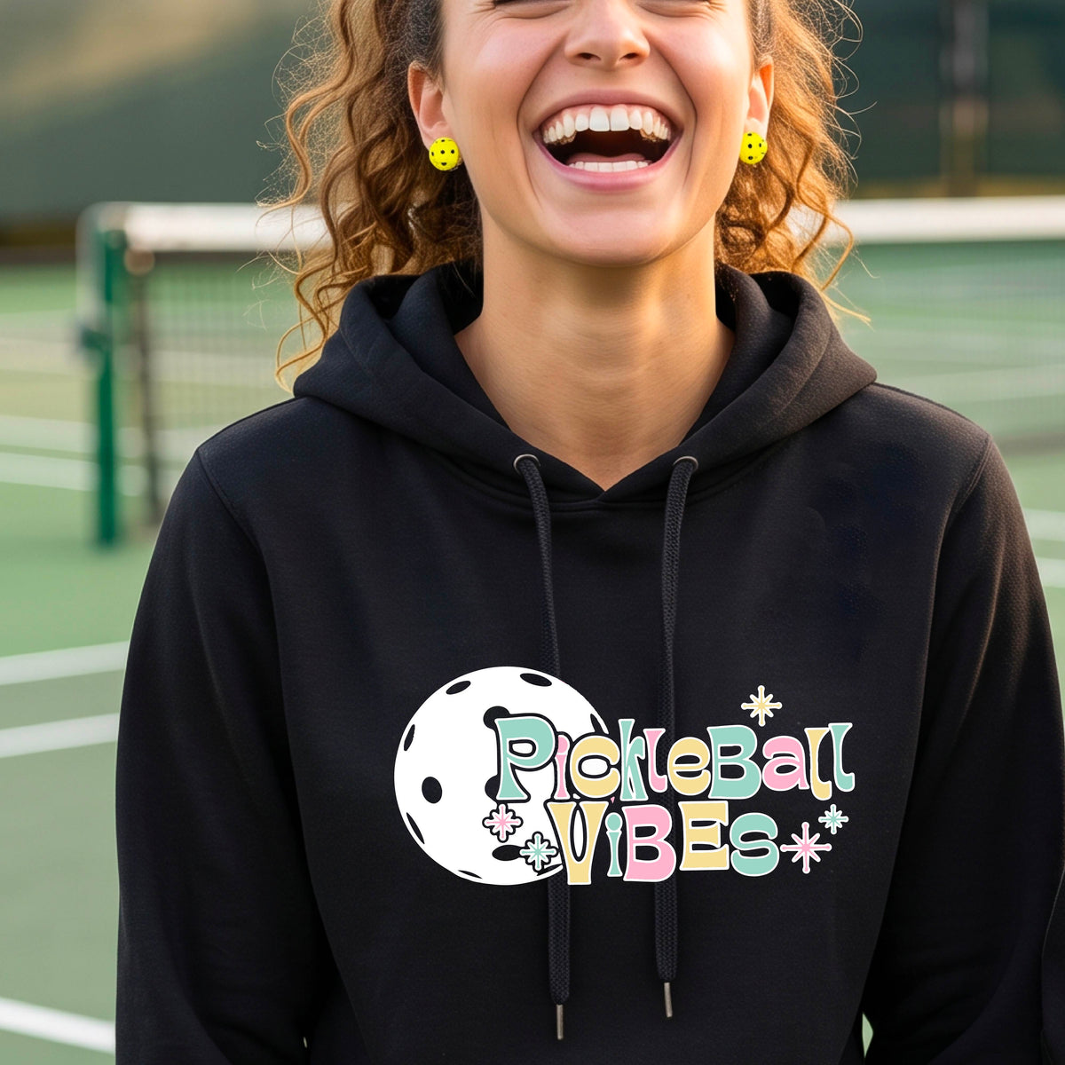 A close-up of a woman on a pickleball court wearing a black fitted hoodie pullover, with a colorful retro style font 'Pickleball Vibes' on top of a white pickleball graphic, on the front of the hoodie. 