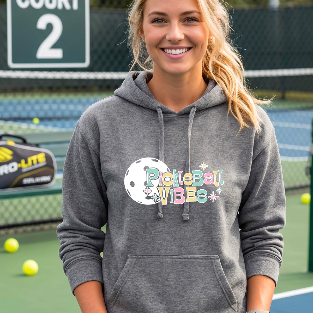A close-up of a woman on a pickleball court wearing a gray fitted hoodie pullover, with a colorful retro style font 'Pickleball Vibes' on top of a white pickleball graphic, on the front of the hoodie. 