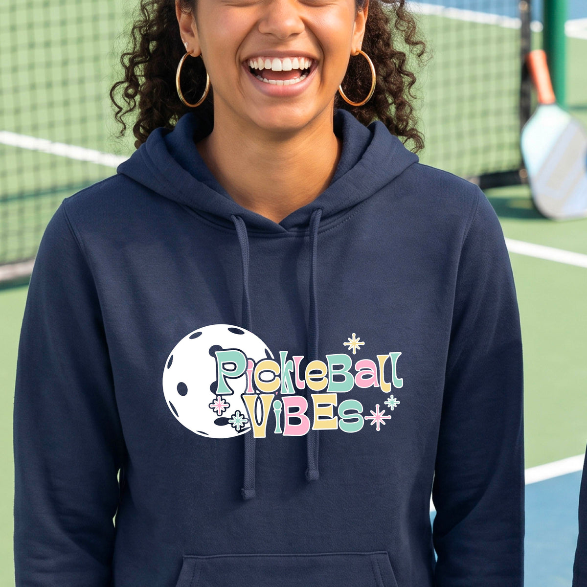A close-up of a woman on a pickleball court wearing a navy blue fitted hoodie pullover, with a colorful retro style font 'Pickleball Vibes' on top of a white pickleball graphic, on the front of the hoodie. 